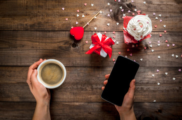 St. Valentine's Day. Woman drinking coffee and watching something on a smartphone, cake with cream and sugar sprinkles (hearts), gift and decoration in form of heart are on table.Top view, copy space
