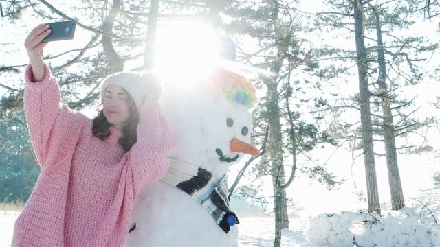 cute girl making photo with a snowman, winter selfie, mobile phone in hand of a young woman making fun selfie photo in winter forest backlit, snow sculpture, fun entertainment
