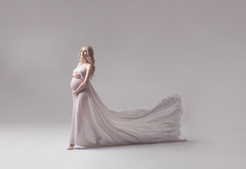 studio portrait of beautiful pregnant young woman in flying white dress with trail on light grey...