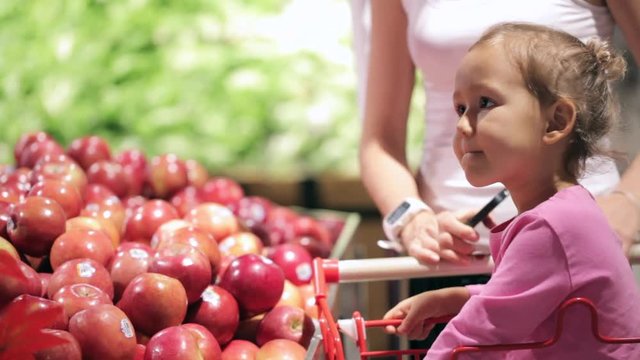 Young mother with little daughter in trolley selecting fruits at supermarket