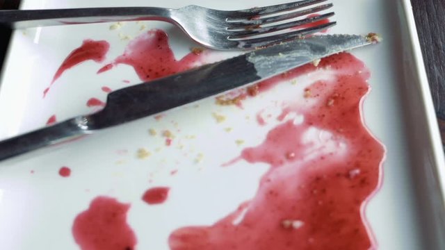 Close-up of empty white plate smeared with pink fruit syrup and crumbs, knife and fork placed on it. Remains of dessert. Detailed shot.