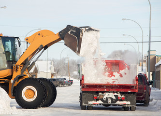 A loader dumping a bucket of snow onto the box of flatbed truck on a main street of small town in winter