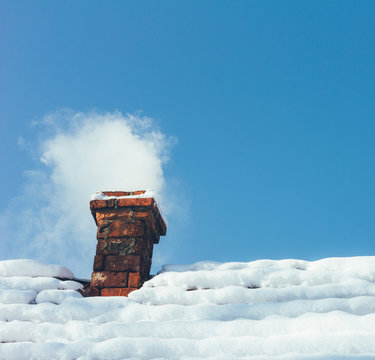 smoke out of a brick chimney on a snowy rooftop home on the background of blue sky