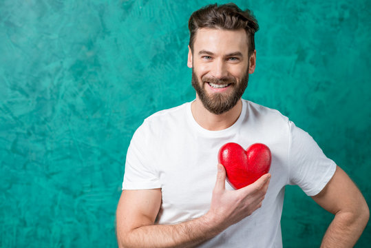 Handsome man in the white t-shirt holding red heart on the painted green wall background. Valentine's Day concept