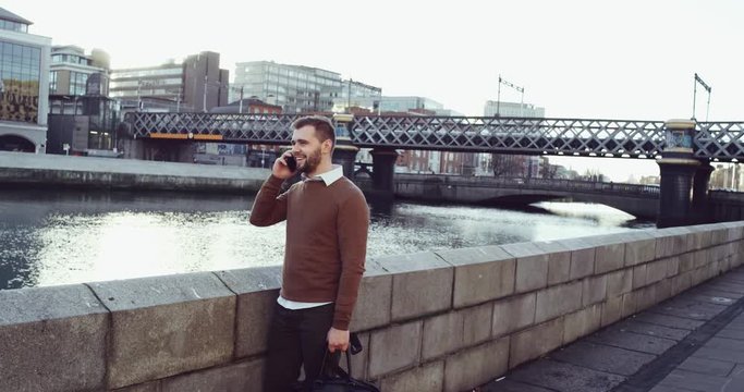 Attractive European businessman using smart phone in front of Drogheda City. Handsome young businessman communicating on smartphone smiling confident near beautiful river . Shot on Red Epic 4k.