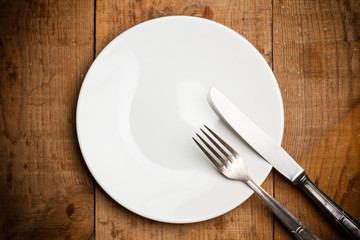 Plato blanco redondo vacío con cubiertos de metal antiguos sobre mesa de madera rústica. Vista cenital