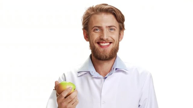 Strong Caucasian young doctor with light beard and medical overall eating apple smiling on white background