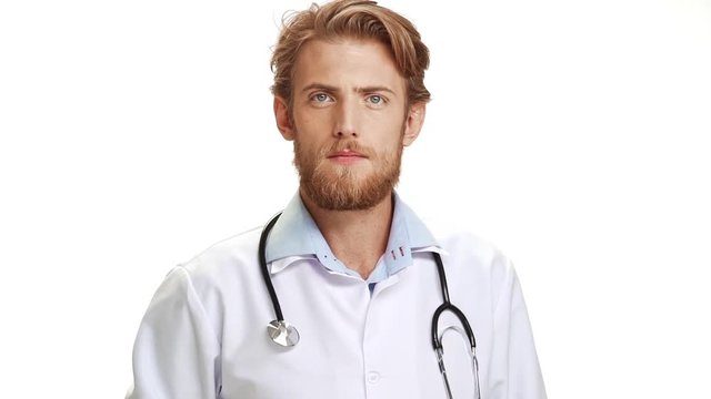 Severe young Caucasian male doctor with light beard wearing medical overall showing fist at camera on white background in slowmotion