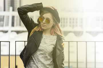 Portrait of young woman in hipster glasses,pot hat and black leather jacket standing and looking forward in city