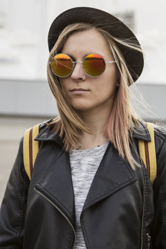 Portrait of young woman in hipster glasses,pot hat and black leather jacket standing and looking forward in city