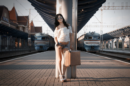 a lonely young brunette woman with an old suitcase is standing on the station platform