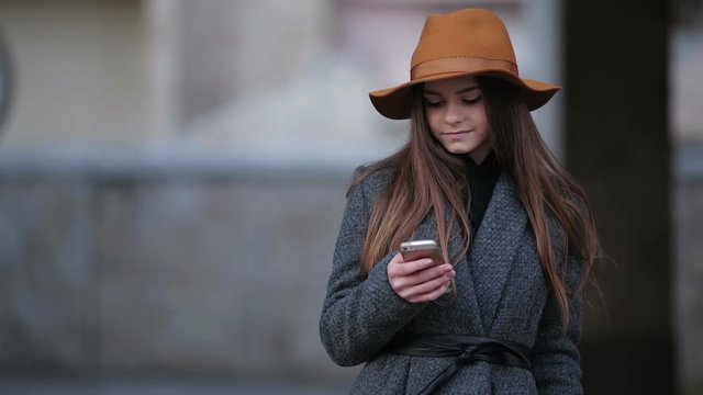 Young Beautiful Girl In A Brown Hat And Coat Uses A Smartphone