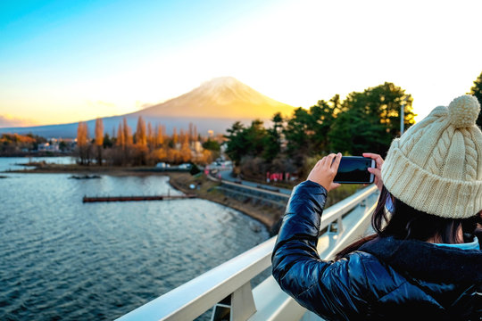 Young Woman taking photo with her phone of the view Mt.Fuji with Beautiful sunset at Lake Kawaguchiko in winter , Japan