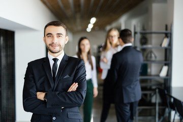 Young manager in front of his coworkers with crossed hands in modern office.