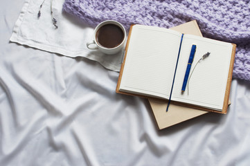 Notebook and cup of coffee with a book and a blanket of lavender on white textiles in bed