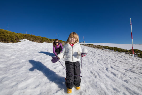 Three years old blonde child with snowball in hand looking, next to woman mother, with white and purple coats on snow in winter mountain
