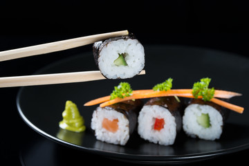 Sticks holding roll with cucumber on the background of fresh rolls with salmon, cucumber and pepper on a plate, served with carrot slices and parsley, black background