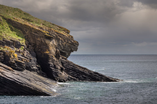Lybster, Scotland - June 4, 2012: Huge brown-gray cliff with green meadow on top protects the north flank of Lybster harbor. It descends slanted into the blue-gray sea under brown-gray skies.