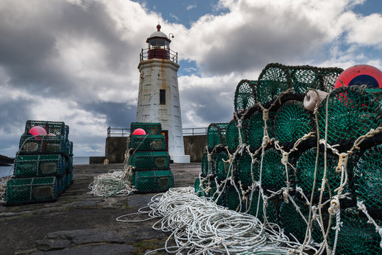 Lybster, Scotland - June 4, 2012: Green lobster traps are stacked on black stone pier of Lybster harbor with white lighthouse in back. White cords and red buoys. Dark clouds in blue sky.