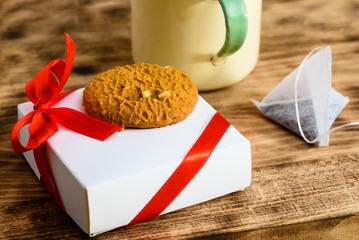 Fototapeta na wymiar Small white present with red bow and cookie on top together with partial teacup and teabag on wooden tray. Focus on cookie
