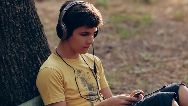 A teenage boy sitting in a park listening to music. Boy with headphones outdoors