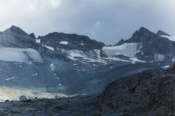 Late afternoon sunlight hits the side of a granite mountain under grey clouds. Two tiny hikers are visible on the ridge line