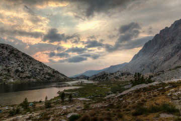 Smoke from a forest fire creates haze and a colorful sunset in an alpine valley with a lake and pine trees