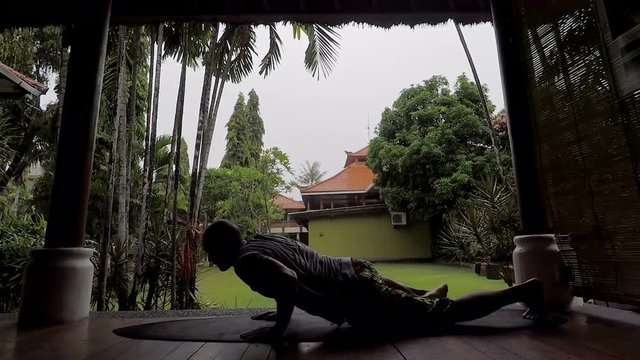 Silhouette of young fit man having training of trunk curl on surfboard on terrace of tropical bamboo house. Athlete trainee is lying down on the ground and then rising in position of riding a board.