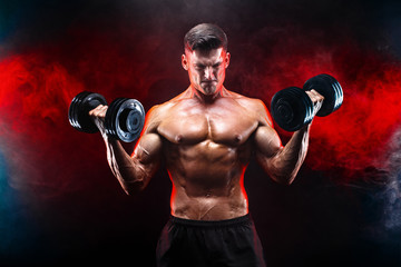 Handsome bodybuilder doing exercise on biceps with dumbbell. Studio shot. Smoke.