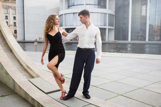 two young people - a man in white shirt and a woman wearing black dress - dancing outside on city embankment against modern glass buildings