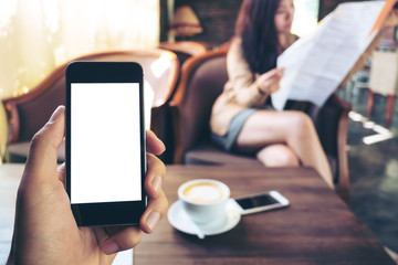 Mockup image of hand holding black mobile phone with blank white screen in cafe and blur asian woman reading newspaper