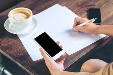Mockup image of hands that writing on papers and white mobile phone with blank black screen , hot coffee and vintage wooden table background