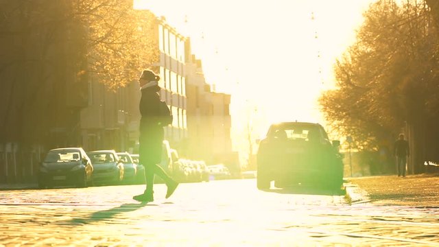 young travel woman in long black coat crossing paved road in the center of town with parked cars on both sides, road, trees and buildings are shining gold under the blind sunlight