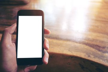 Mockup image of hands holding black mobile phone with blank white screen and vintage wood table background