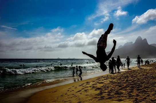 Silhouette of the man performing back somersault at Ipanema beach with silhouettes of swimming and walking people, blue sky, white clouds and Dois Irmaos Mountain at background, Rio de Janeiro, Brazil