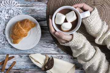 Hand holding cup of hot chocolate and marshmallows with croissant, top view