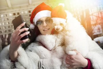 Beautiful young woman with her dog. Wearing Christmas hats. Smiling young woman holding her dog while taking a selfie. Lens flare in a background.