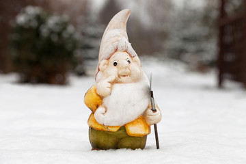 Garden dwarf with shovel in winter garden. Garden gnome on snow in yard. Ornament figurine, winter. Gnome in snow-covered curtis. Garden with fairy statuette. Troll with shovel in snowy garth
