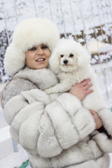 Winter beauty. Smiling young woman in the wintertime with her dog. Woman wearing white winter fur coat while holding her dog. Christmas decorations in the background.