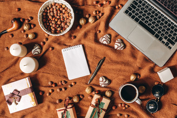 Home workspace in romantic style with laptop, notebook, heart shaped biscuit, pen, coffee cup, gifts, nuts on brown fabric cover. Empty sheet in center. Copyspace, top view, flat lay