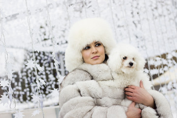 Winter beauty. Close up of a smiling young woman in the wintertime with her dog. Woman wearing white winter fur coat while holding her dog. Christmas decorations in the background.