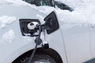 Charging an electric car in winter time, automobile covered by fresh snow, electric cord plugged in the car