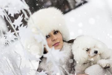 It is snowing! Beautiful woman in the winter time with her dog. Woman wearing a winter coat and a fur hat. Snowfall around her.