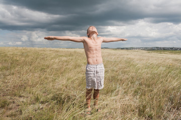 young boy stands in a field. boy in shorts. boy stands in the desert. feather in the field. boy looking up at the sky. dreamer