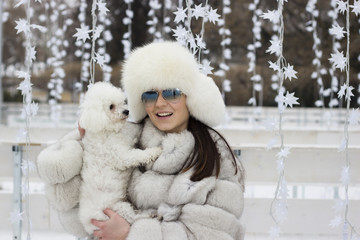 Obraz premium Beautiful woman playing with her dog outdoors. Winter time. Playing with the dog at the park. Woman wearing a white fur coat. Christmas decorations in the background.