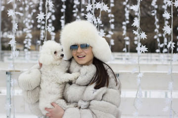Beautiful woman playing with her dog outdoors. Winter time. Playing with the dog at the park. Woman wearing a white fur coat. Christmas decorations in the background.
