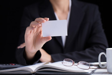 A business woman in black suit holding white blank business card in her hand on a working desk.