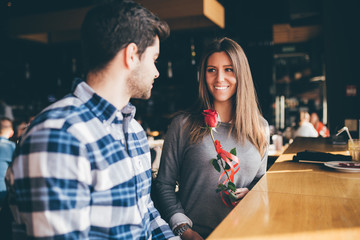 Beautiful young couple in love enjoying together at cafeteria, and man is giving a red roes as surprise to his girlfriend. 
