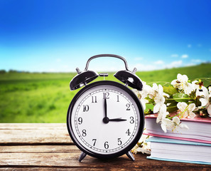 Alarm clock with books and spring blooming branch on wooden table against landscape background. Time change concept