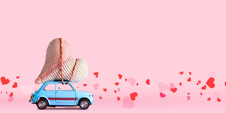 Blue retro toy car delivering craft heart for Valentine's day on pink background with confetti